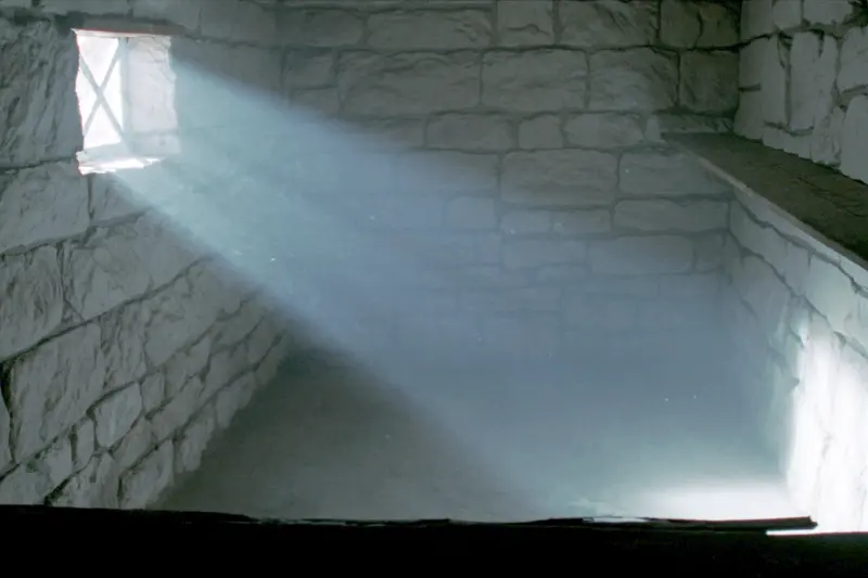 A whitewashed fieldstone cellar with one small window throwing a beam of daylight across the floor and a wooden shelf on the wall