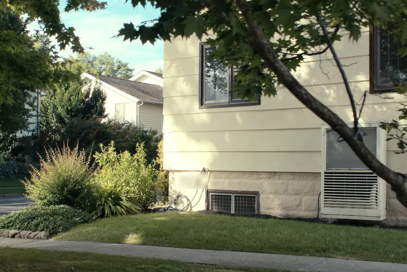 An air conditioning condenser beside cream siding in summer, hostas and a coiled garden hose at the foundation, maples overhead
