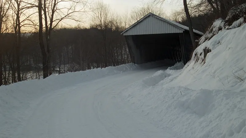 A white covered wooden bridge at the bend of a snowy road, bare trees and a low winter sun behind it