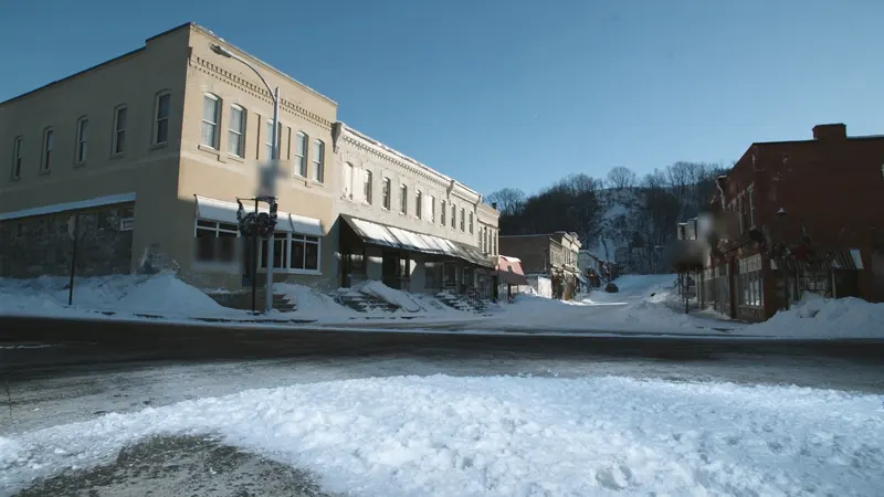 A snowy small town main street of cream brick and limestone storefronts under a clear blue winter sky, plowed snow along the curb