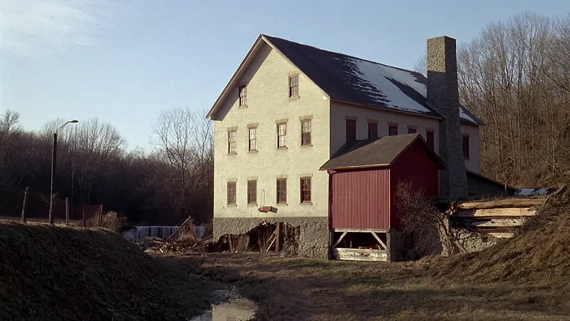 A tall cream brick mill with a stone chimney and a red wooden shed beside a small dam, bare trees behind, winter sun