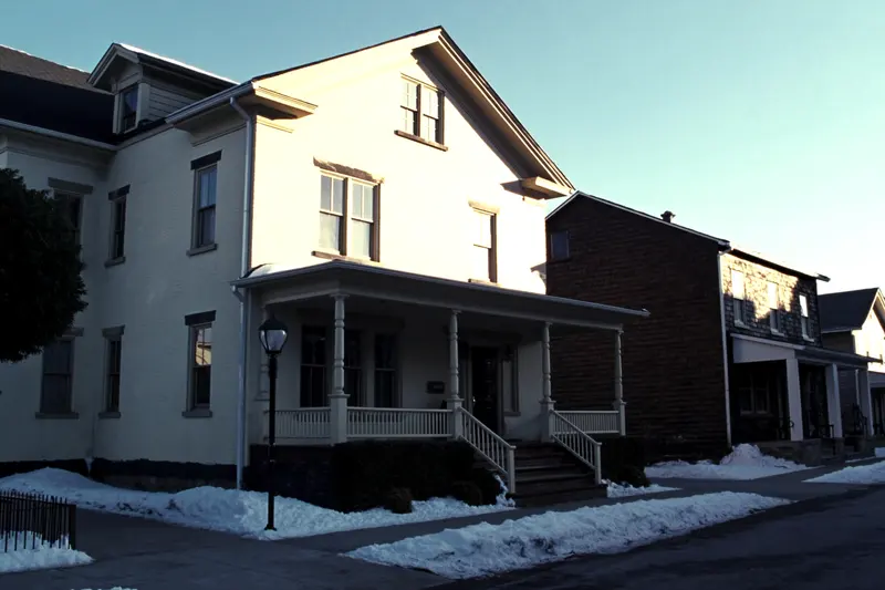 A cream painted brick house with a deep front porch and a street lamp, lit by low winter sun, snow along the sidewalk